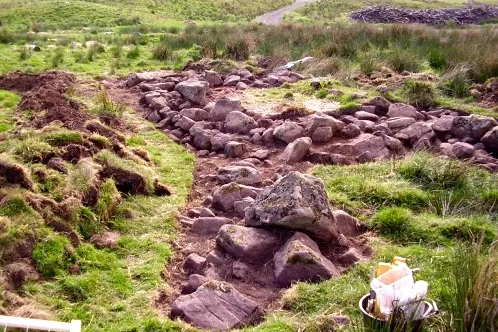 Excavation of a medieval cruck-framed building, with substantial stone footings exposed in the turf. The scale of the rubble suggests a longhouse or byre of considerable size.